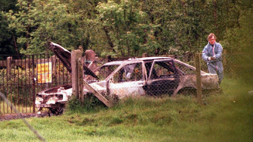 A man in a forensic boiler suit is standing at the rear of a burned out car. The bonnet and back door of the car are opened. The vehicle has been destroyed by fire so no longer has any paint, windows or tyres. It is parked in an area surrounded by security and farming fences with lots of grass. There are also trees and a danger sign in the background.