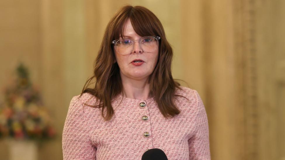 Caoimhe Archibald is speaking into a mic. She has transparent glasses on and her hair has been styled in waves. She's wearing a pink jacket with gold buttons.