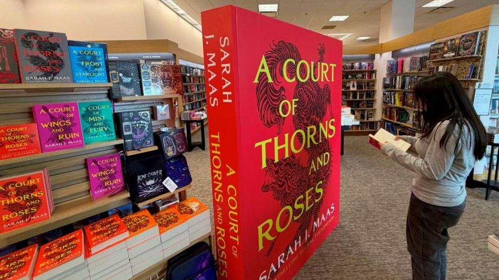 Women wearing grey and black standing in front of large ACOTAR series display.
