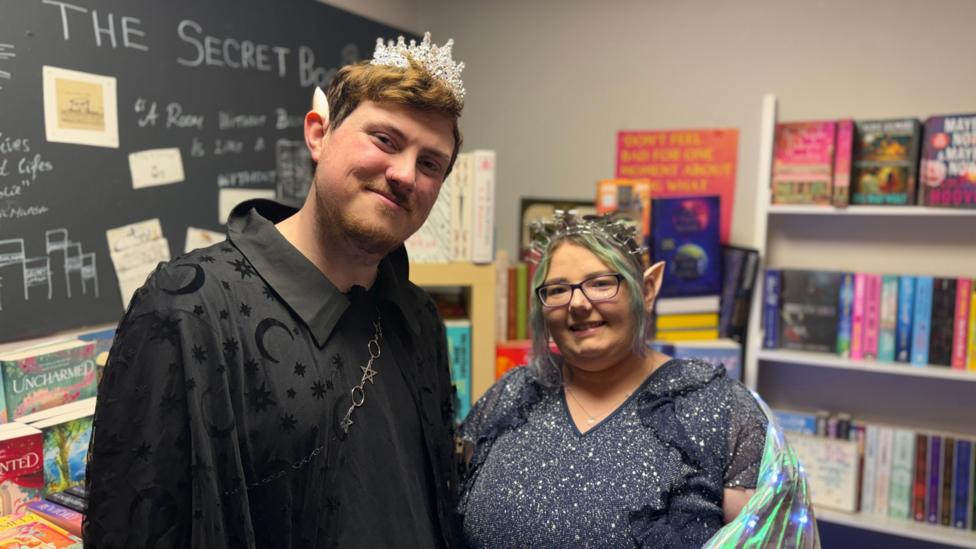 David and Sami are dressed up in costumes for the ACOTAR event. David on the left is wearing an all-black attire, whereas Sami on the right is wearing a glittery blue dress with light up wings. They are both smiling at the camera while standing in front of a stacked bookshelf.