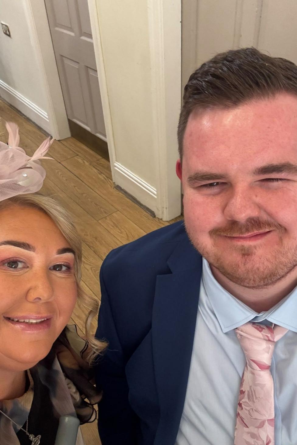 A woman and man looking at the camera. The man is on the right and has a dark blue suit jacket, light blue shirt and pink tie. He has short brown hair. The woman is on the left and has a pink fascinater in her hair.