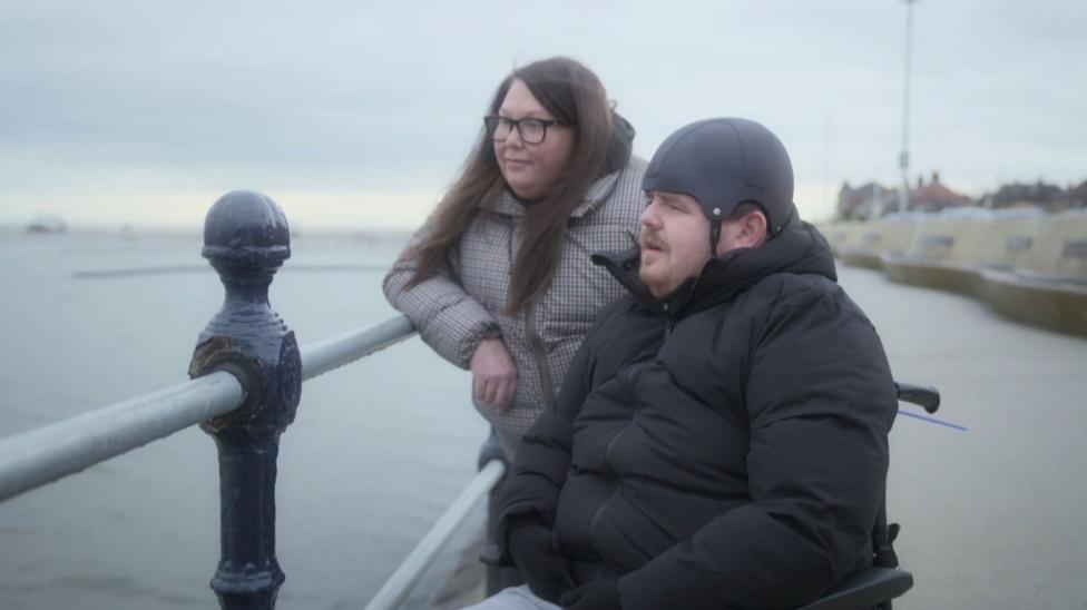 A man in a black puffer coat in a wheelchair with a black helmet on. He is looking out to sea. There is a woman next to him with long dark brown hair and a checkered puffer coar.