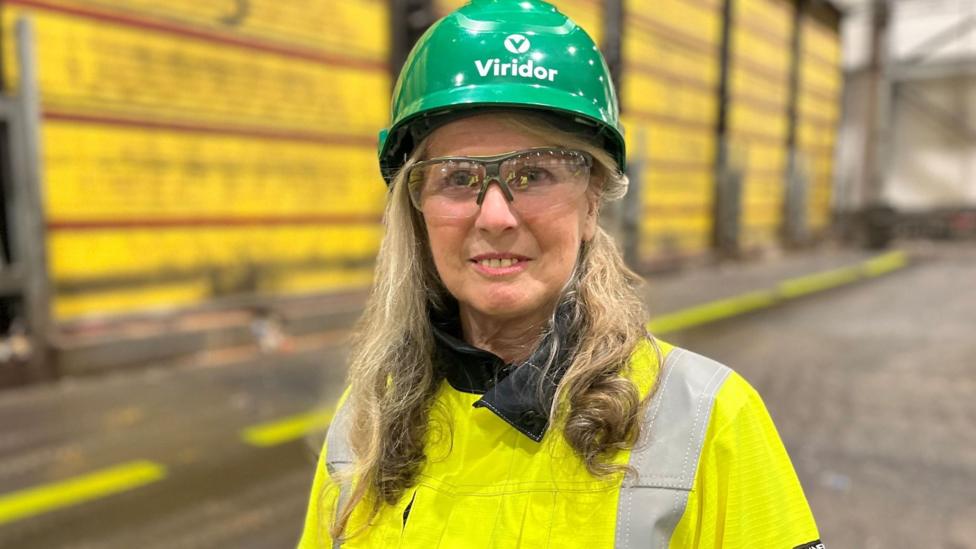 A woman in a yellow high-vis jacket, goggles and a green branded helmet standing in front of yellow roller doors.