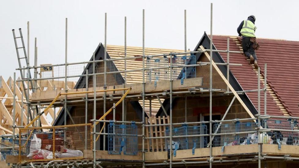 There are three new housing being built. The one on the right has red slate times on top of it, with a man in hi-vis putting them on. In the middle, there is a house that has its wooden panels and bricks but needs the slate on top. On the left there is a frame of a house. In front of it is scaffolding. 