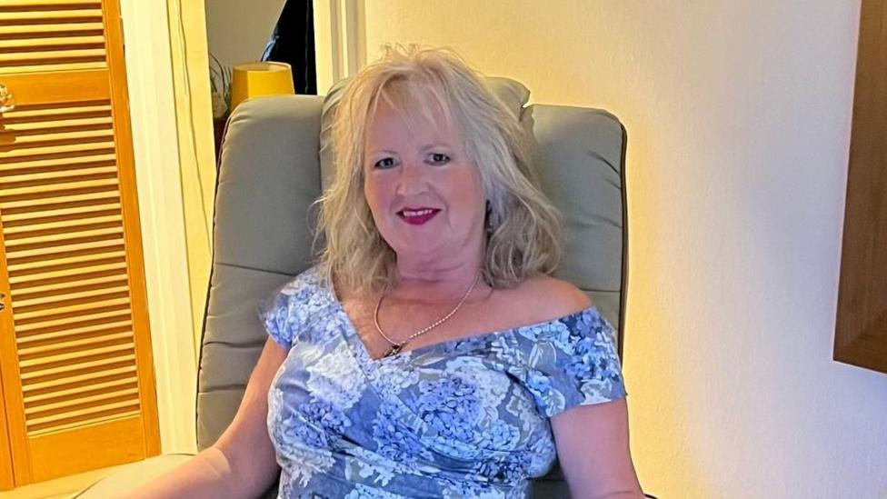 There is a woman in a blue dress, a silver pendant and blonde hair. She is sat down in a beige chair in front of a doorway.