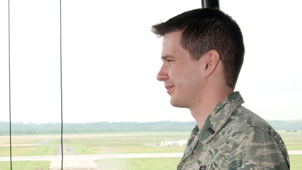 A man in a US Air Force uniform stands in front of a window looking out onto a airfield.