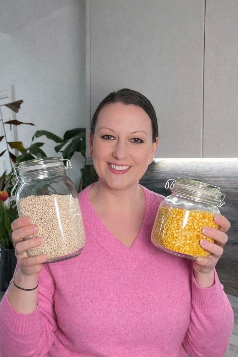 Ana stands in a kitchen holding two large glass storage jars. One jar contains small pale grains, while the other contains yellow dried pulses. The person is wearing a bright pink jumper. Kitchen cupboards and houseplants can be seen in the background, and the image is brightly lit with natural-looking light.