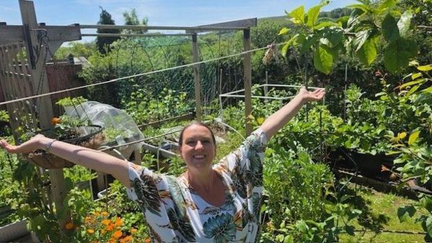 Ana stands in the middle of a lush garden with both arms stretched wide. She is wearing a loose shirt with flowers and leaves on it and her brown hair tied up. The garden is densely packed with vegetables, flowers and greenery, including raised beds, climbing plants and fruit trees. Bright orange flowers bloom in the foreground, while a wooden frame and netting structures for growing plants stand behind. The photo is taken on a sunny day, with blue sky visible above and rolling green hills in the distance.