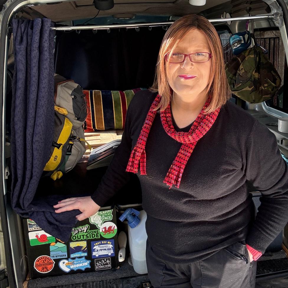 A woman with shoulder length brown hair is standing at the open back of a van that has been converted for practical, outdoor use. Inside the van are various items including backpacks, a striped blanket, hanging curtains and multiple stickers covering a small storage unit. The woman is wearing a black top and a red patterned scarf, resting one hand on the edge of the van while the other is in a pocket. Sunlight is shining into the van, highlighting the gear and interior setup.