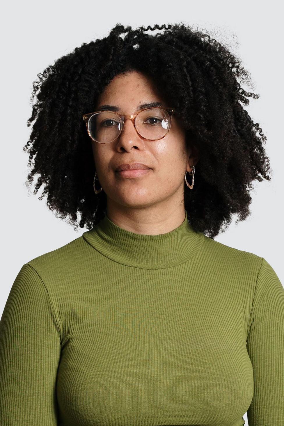 A studio-style head and shoulders portrait of Yasmin Ibison standing against a plain light-grey background. She is wearing round glasses, hoop earrings and an olive-green high-neck top. 