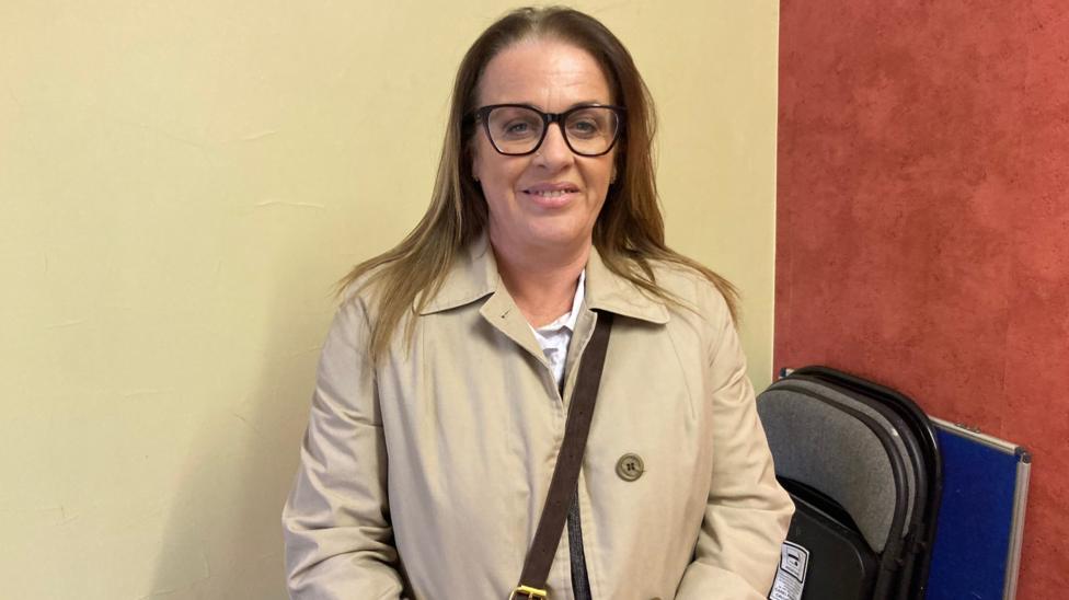 A person wearing a beige trench-style coat and dark-framed glasses stands indoors against a two-tone wall painted cream and red. A brown cross-body bag strap is visible across the front of the coat. In the background, several black and grey chairs are stacked upright beside a blue panel.