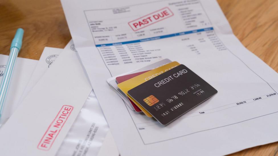 Credit cards are placed on top of household bills spread across a wooden table. One document is marked "PAST DUE" in large red lettering, while a nearby envelope is stamped "FINAL NOTICE". Additional envelopes and a blue pen are placed beside the paperwork.