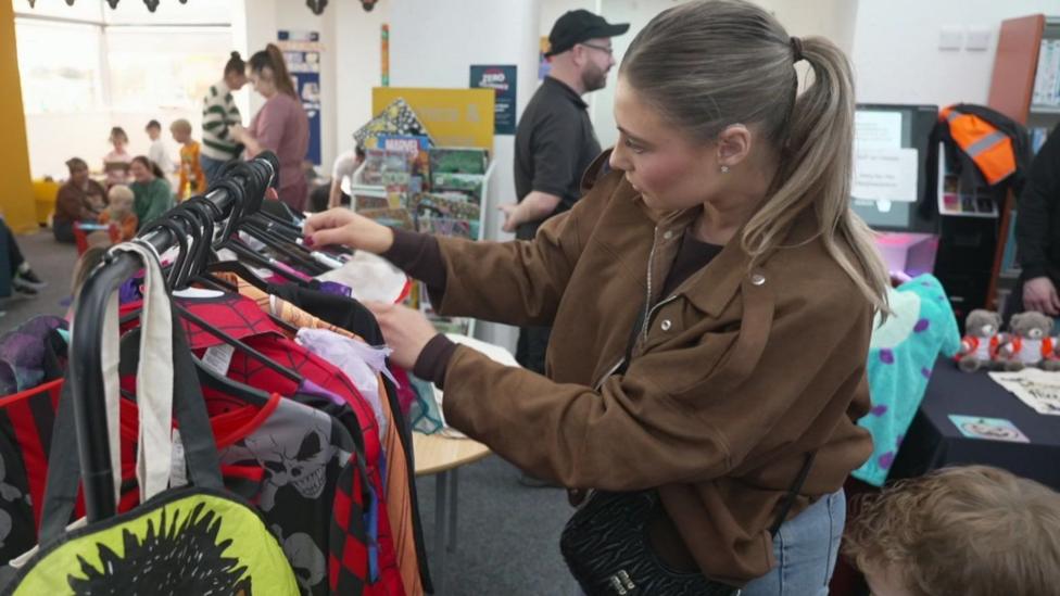 Woman with light brown hair tied in a ponytail, wearing a brown jacket, looks through a rail of donated Halloween costumes at Prescot Library. Other adults and children are gathered in the library, with bookshelves and a display of comics visible behind them.