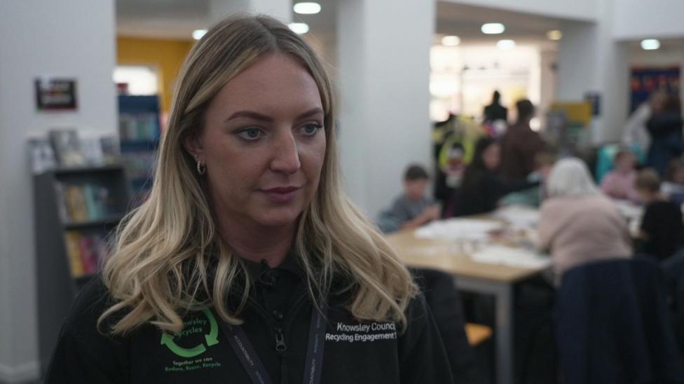Barlow, from Knowsley Council's recycling engagement team, is pictured inside Prescot Library. She has long, wavy blonde hair and is wearing a black council uniform with a green Knowsley Recycles logo. Other adults and children are gathered in the library, with bookshelves and a display of comics visible behind them.