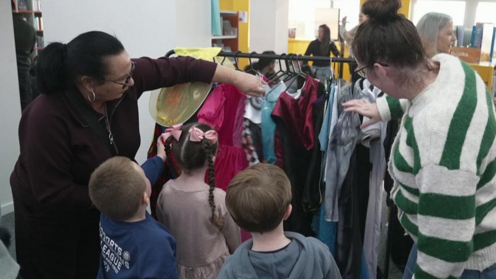 Two women and three children browse a clothes rail filled with colourful Halloween costumes at Prescot Library.
