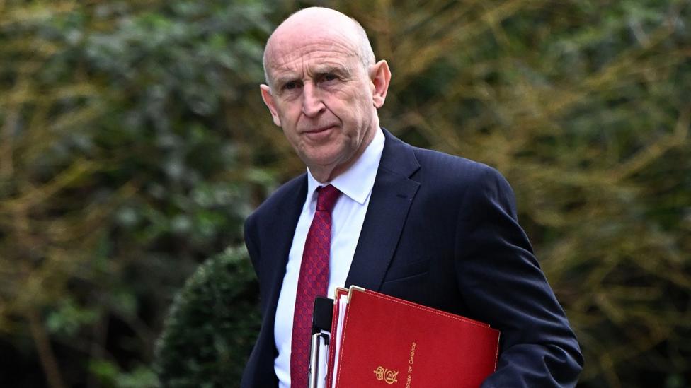John Healey, a man with shaved head, wearing a navy suit, white shirt and claret tie. He is carrying a red book.