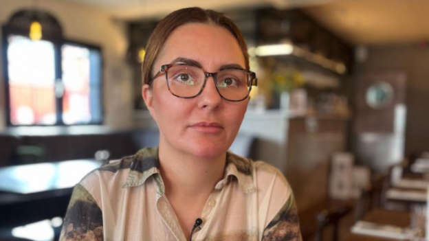 Christina Taylor, a woman with brown hair and glasses, who is wearing a beige top with green and brown splashes of colour.