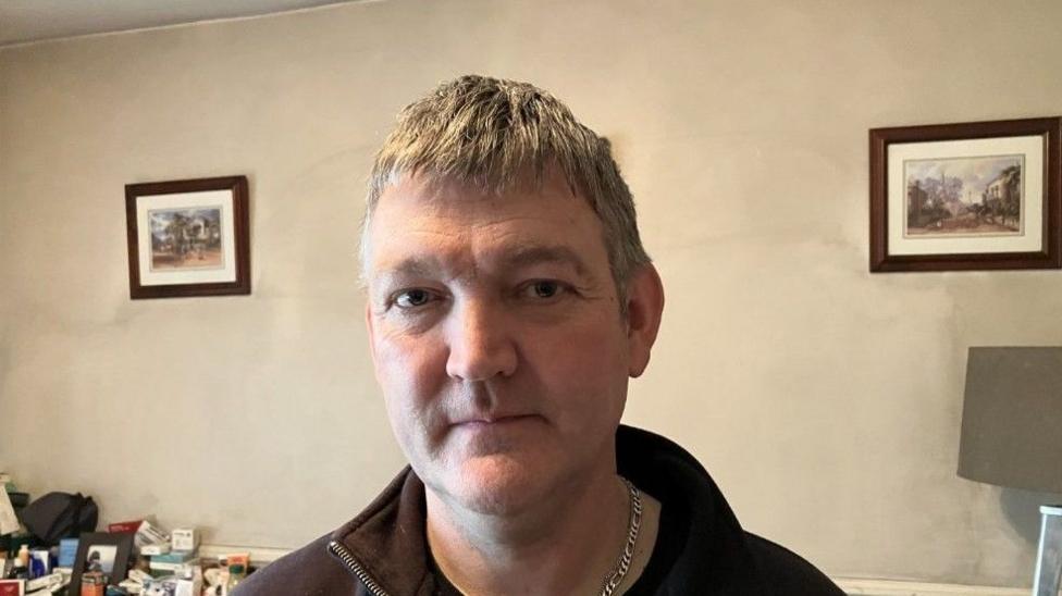 Derek Moore, a man with grey hair, wearing a brown top and a silver necklace. There are two pictures on the wall in the background.