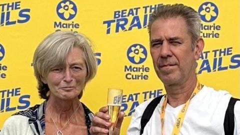 A woman stood next to a man who has completed a race for Marie Curie. He has a medal around his neck and holding a glass of bubbly.