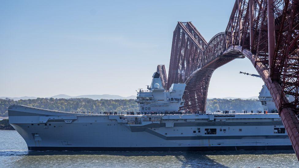 A large warship passes under a red stell bridge