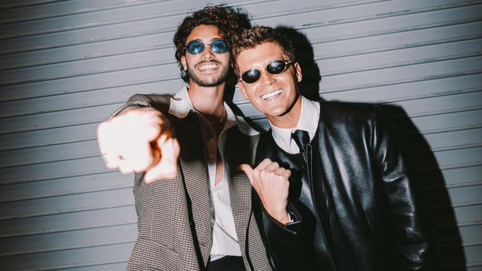 Two men stood next to each other. One is pointing at the camera wearing a suit jacket, open buttoned shirt and blue tinted glasses. The other wears a leather jacket, shirt. tie and black tinted glasses.