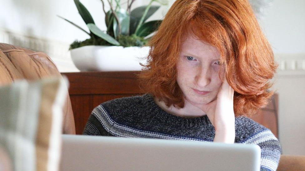 A young woman with red hair looking at a laptop with a serious and thoughtful expression