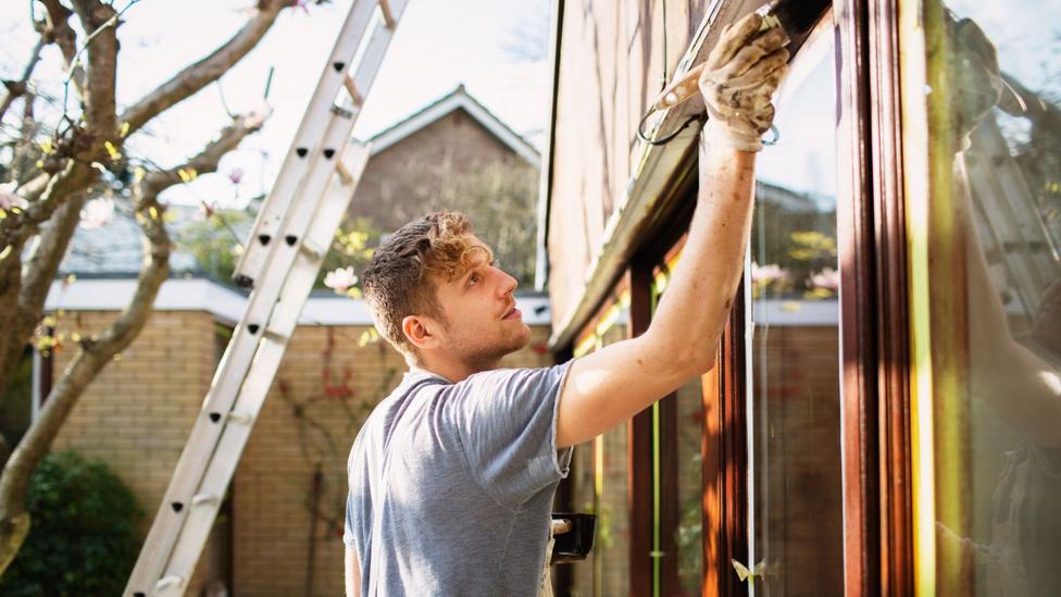 A young man at work painting the outside of a residential property. He is standing on the ground and there is a ladder seen in the background