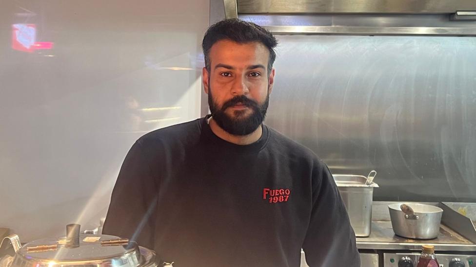 A man stands in a kitchen of a restaurant. He is looking into the camera. He is wearing a black t-shirt with the words 'FUEGO 1987' printed on the chest.