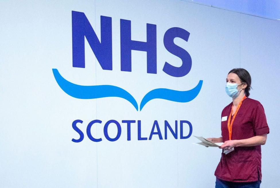 A white wall with blue lettering which says 'NHS Scotland' - a medical professional wearing maroon scrubs and a surgical mask walks by