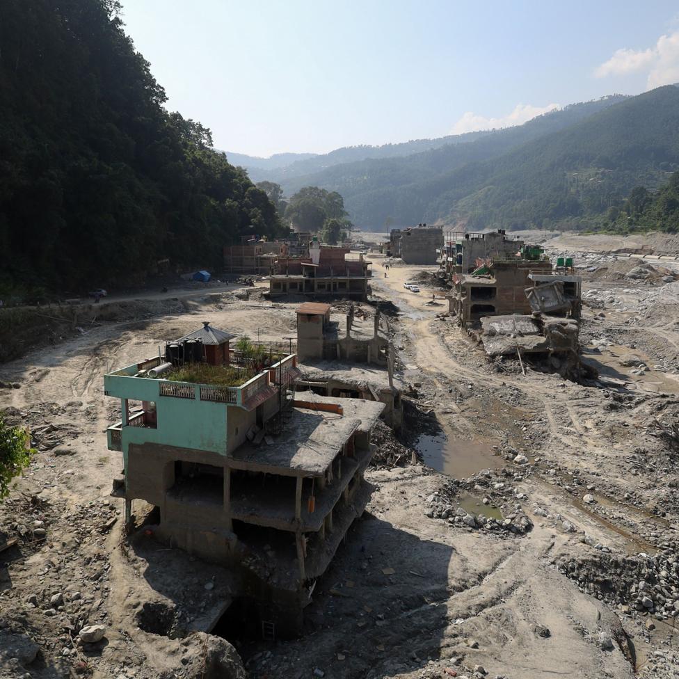 Flood-damaged buildings in a vast landscape of mud, gravel and debris in Trishuli Bazar, Nepal, nearly two months after devastating floods swept through the valley. Several structures are partially collapsed or left stranded above eroded ground, while a widened river channel and scarred floodplain stretch through the settlement between steep, forested hills.