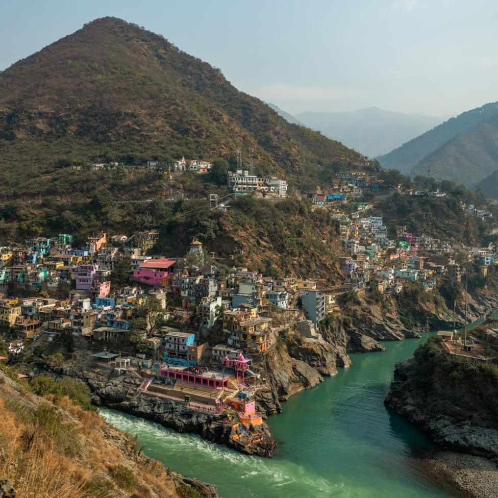 The turquoise waters of two rivers meet at Devprayag in the Indian Himalayan state of Uttarakhand, with colourful buildings clustered on steep rocky hillsides above the confluence. Surrounded by rugged mountains, the rivers wind through a narrow valley lined with temples, homes and roads carved into the slopes.