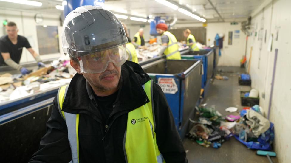 A worker wearing a high-visibility vest, safety helmet and protective goggles stands beside a waste-sorting conveyor belt inside a recycling facility. In the background, several other workers in high-visibility clothing sort mixed waste by hand along the conveyor line. Overhead fluorescent lights illuminate the industrial workspace, where piles of recyclable materials and waste are visible on the conveyor and on the floor.
