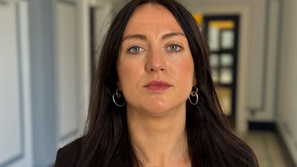 A woman with long brown hair, wearing a black blazer and gold hoop earrings... Stood in an out of focus hallway. She has a neutral expression.