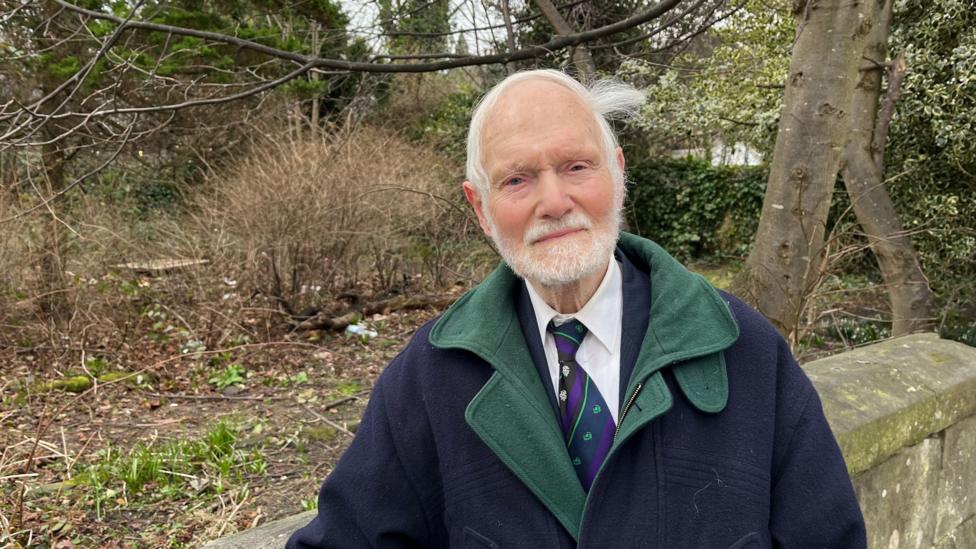 A man with white hair and a beard, wearing a blue and green coat over a white shirt and purple tie. He looks into the camera with a neutral expression, while standing in front of an overgrown patch of land behind a stone wall.