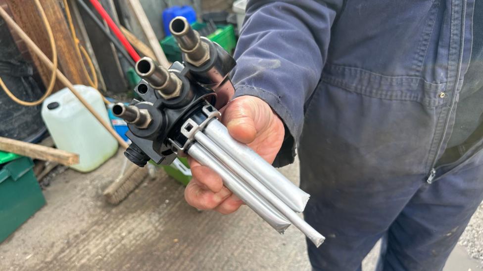 A close-up of a fuel sending unit. The pipes have been cut. A man is holding it.