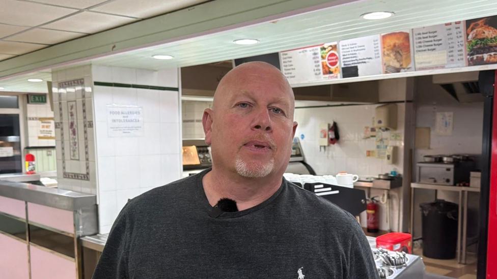 A bald man with a grey goatee in a dark grey tshirt. He is standing in a chip shop