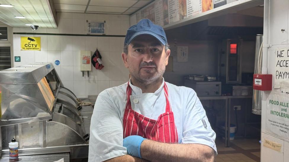 A man in a navy blue cap, white chef unifrom and red apron. He has his arms crossed and is standing behind the counter in a fish and chip shop