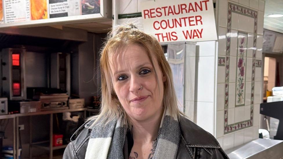 A woman with blonde hair and a grey scarf is looking at the camera