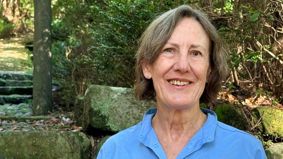 Diane Tincher smiles at the camera. She is wearing a blue shirt and standing in her garden