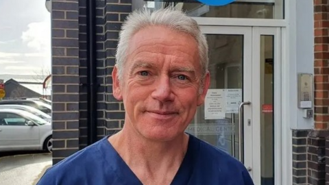 Shows a man with grey hair in a blue v-neck top standing in front of a building and cars to the left