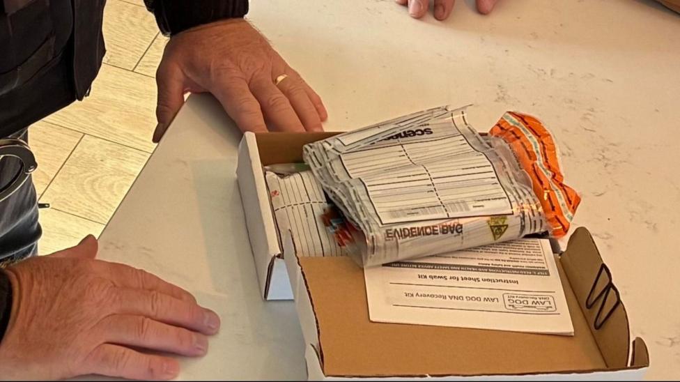 A close up of two people standing by a kitchen table, their heads and upper bodies out of shot. The man on the left is a police officer in uniform, the man on the right is wearing a brown bodywarmer and red sleeved top. On the table between them is a cardboard box with some scientific leaflets and sample bags in it.