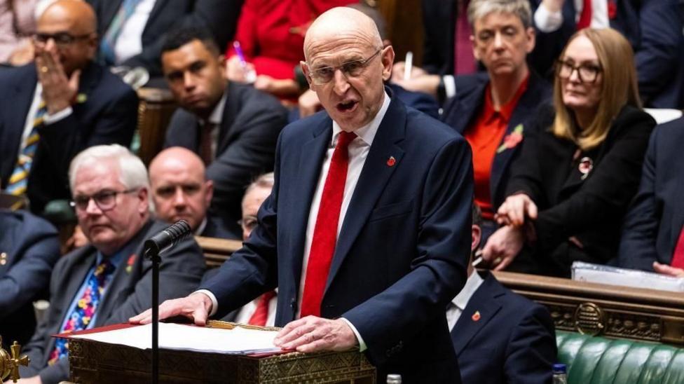 John Healey - a bald man with glasses, a suit and red tie - gives a speech at the dispatch box in the House of Commons as other MPs behind him watch.