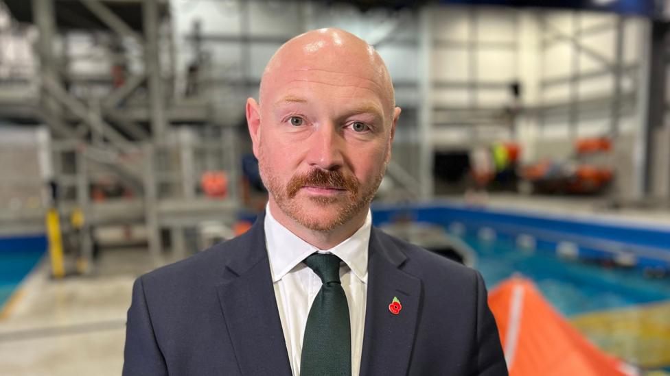 A bald headed man with a moustache, wearing a tie and dark blue suit. A training facility including a pool is in the background