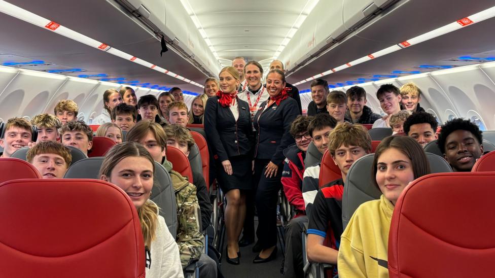 A group of college students sat in passenger seats on a plane. In the middle aisle are airline staff. They are all smiling and looking into the camera. 