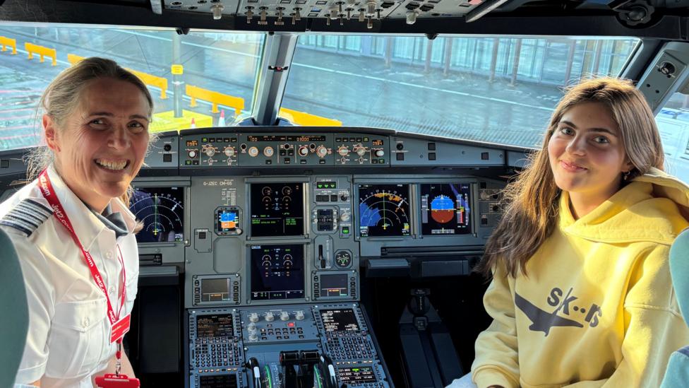 A woman wearing a white pilot's shirt and a Jet2 lanyard and a young woman wearing a yellow hooded top smiling while sitting next to each in a plane's cockpit.