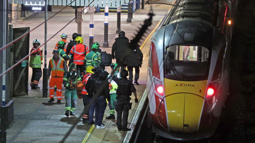 Emergency services personnel, wearing protective gear and many in high-visibility clothing, near an Azuma train. They are standing on the platform to the left of the train, which has its front lights on.