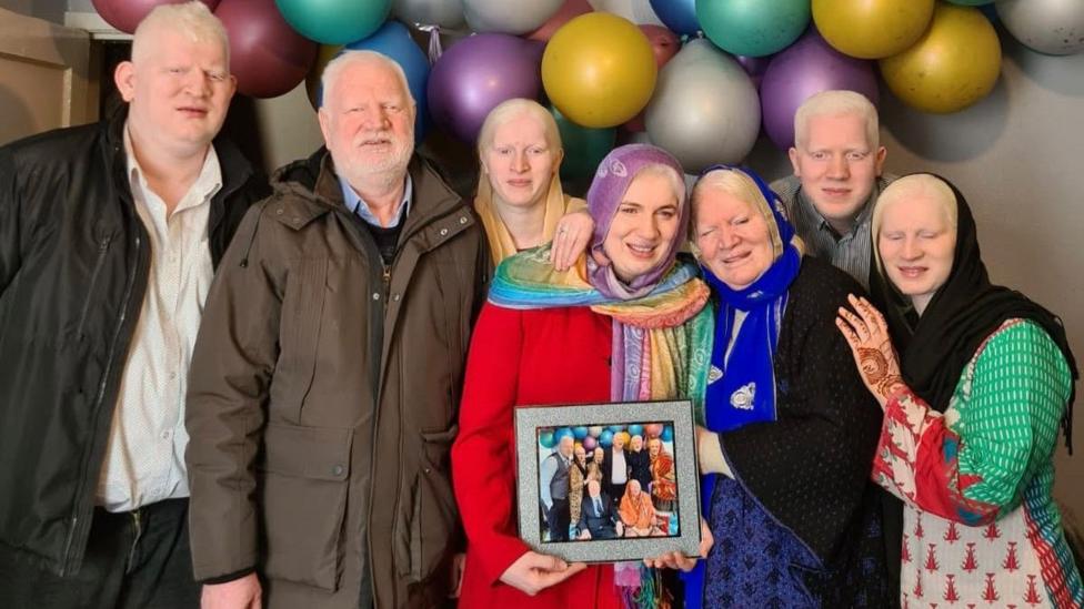 Six siblings, three men and three women, have white hair and eyelashes and are standing around their mother, who also has white hair, as well as a dark blue headscarf on. Behind the group are colourful balloons.