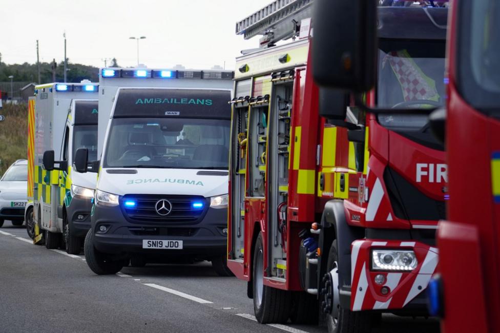 A fire engine with red, yellow and white markings parked on road. Behind it are two white ambulances with their blue lights flashing.