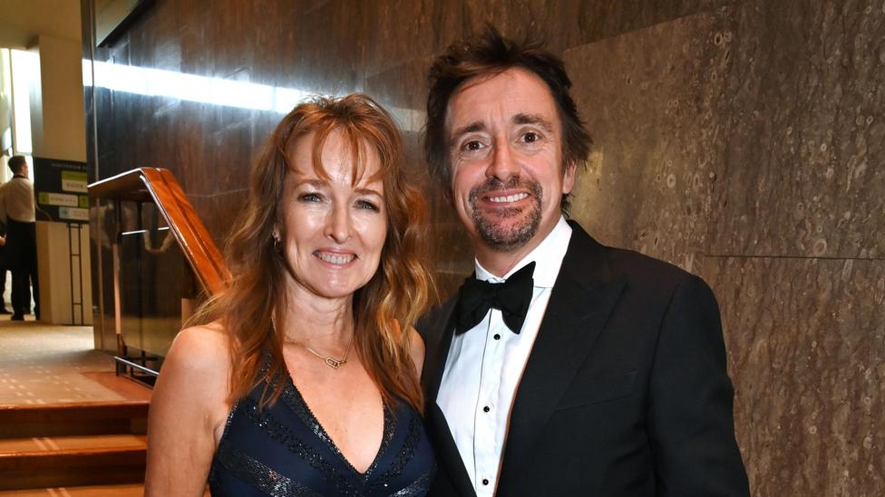 Richard Hammond stood next to his wife Mindy. He is wearing a black tuxedo and she is wearing a black dress, and has wavy red hair.