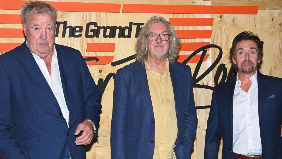 L-R Jeremy Clarkson, James May and Richard Hammond. They are wearing blue suits and standing in front of promotional sign for The Grand Tour TV programme